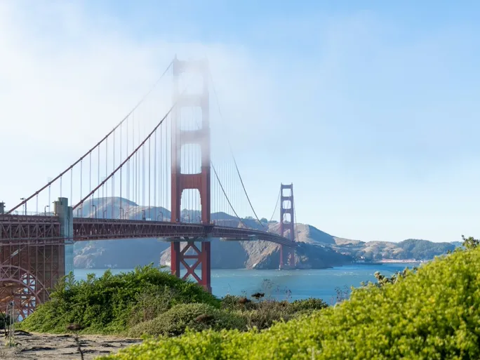 The Golden Gate Bridge spanning San Francisco Bay from headland to headland under a sunny sky, a small wisp of fog capping the near tower.